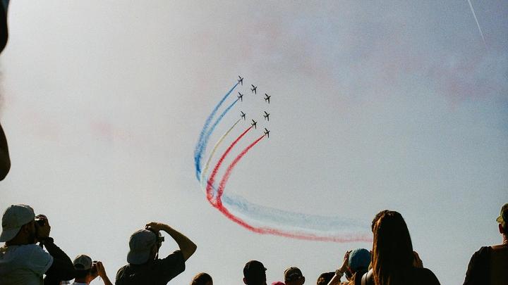 A group of people watch an airshow with several planes flying in formation and leaving red, white, and blue smoke trails in the sky.