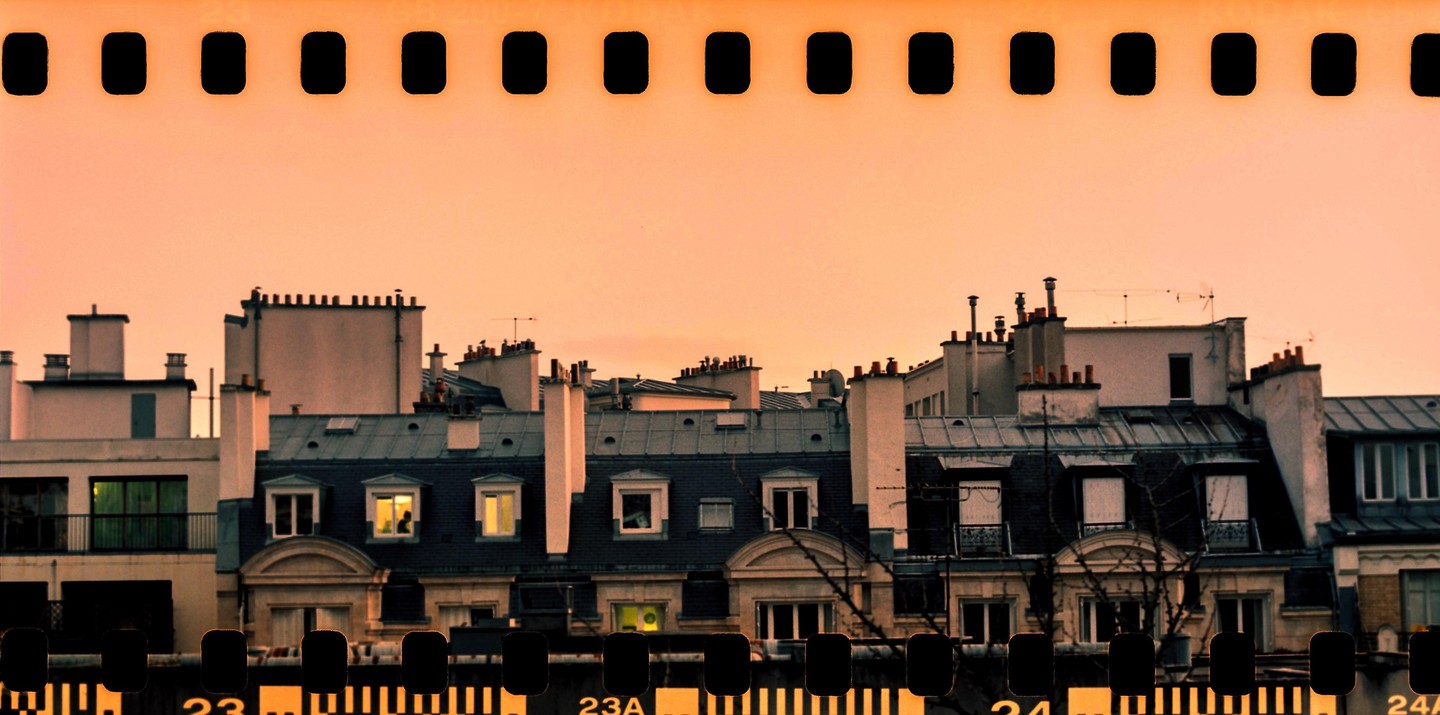 A panoramic view of rooftops with chimneys and windows, some illuminated, is captured under a warm, clear sky.