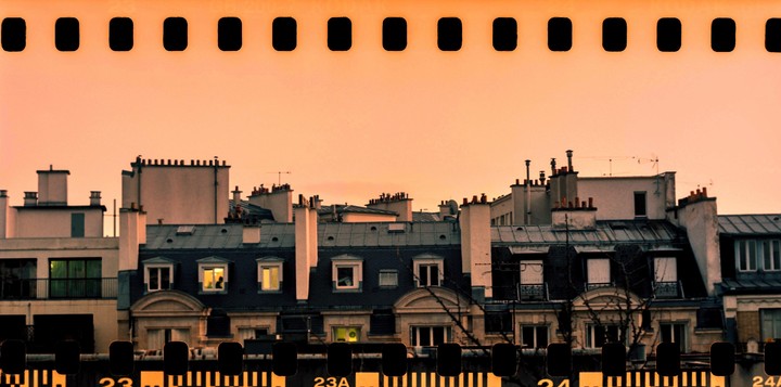 A panoramic view of rooftops with chimneys and windows, some illuminated, is captured under a warm, clear sky.