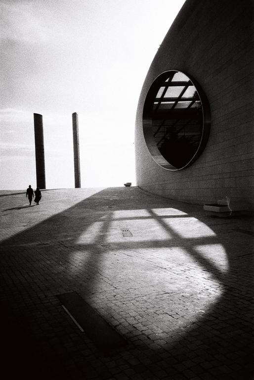 Two people walk on a paved surface next to a modern building with a large circular window, casting a grid shadow.