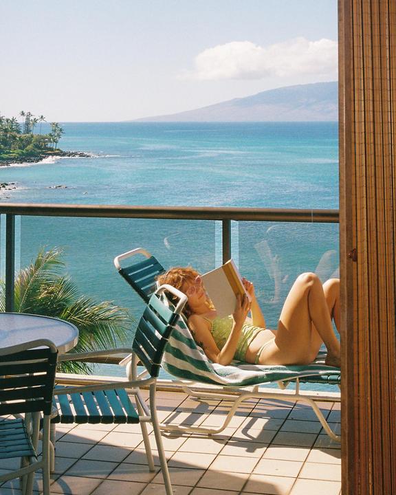 A woman reads a book while relaxing on a lounge chair on a balcony overlooking the ocean.