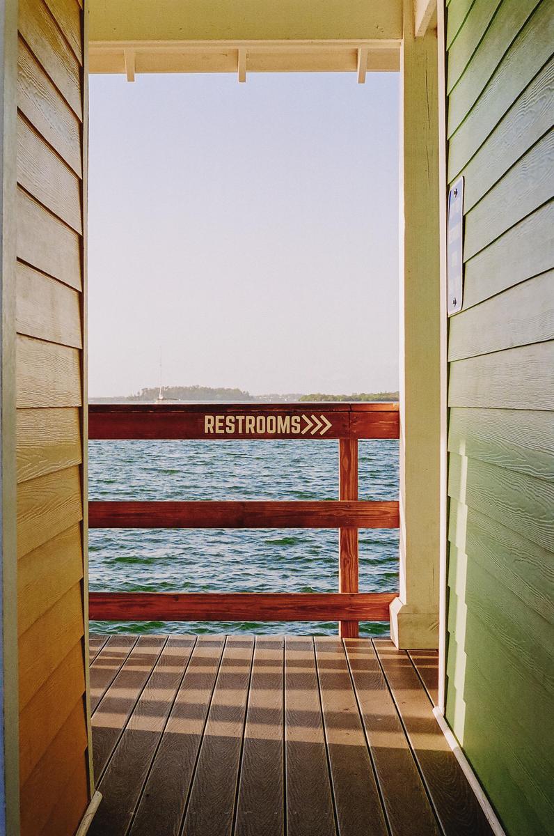 A wooden deck and railing with a "RESTROOMS" sign overlooks a body of water with land in the distance.