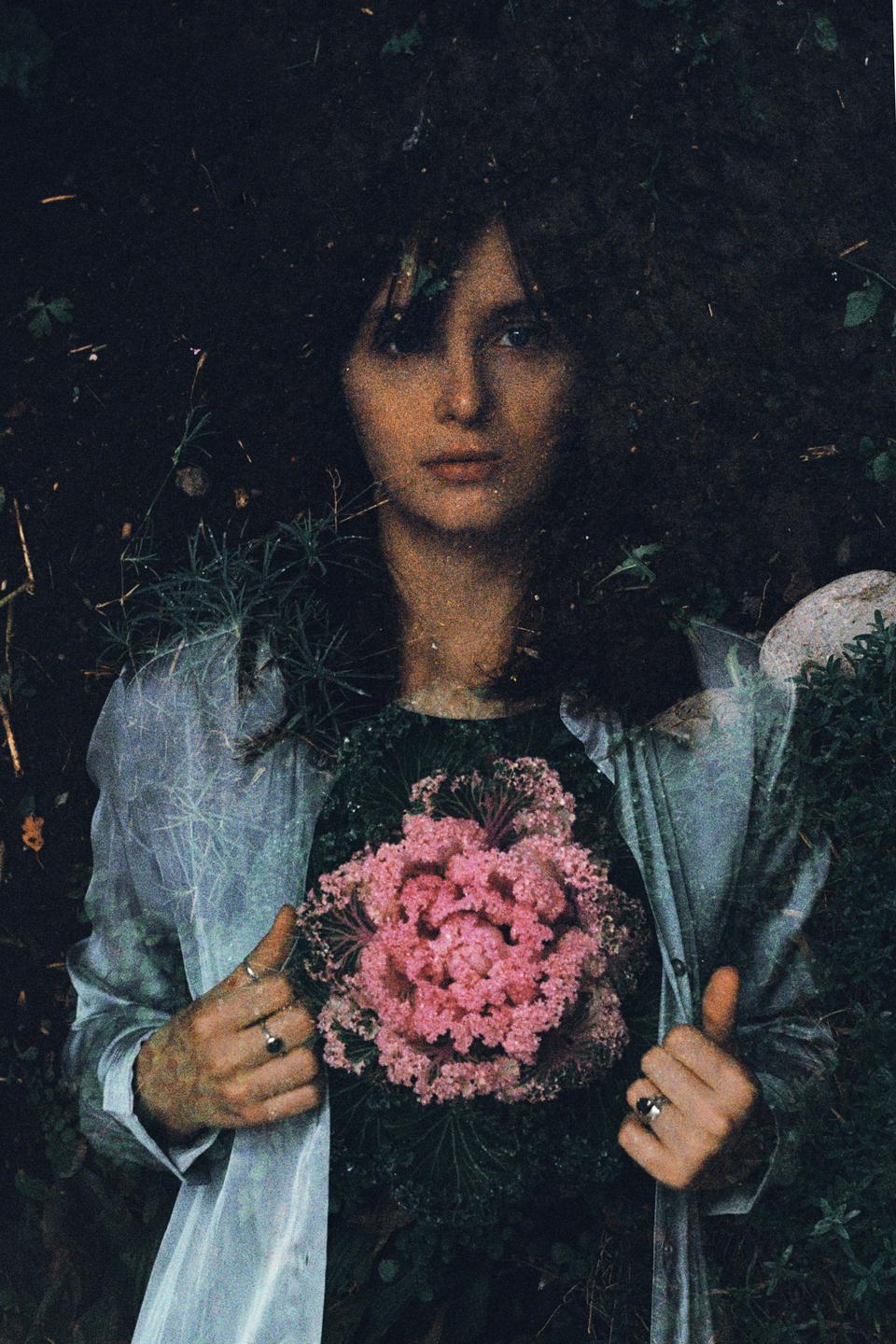 A woman with dark hair and a light shirt holds a large pink ornamental cabbage against a dark, leafy background.