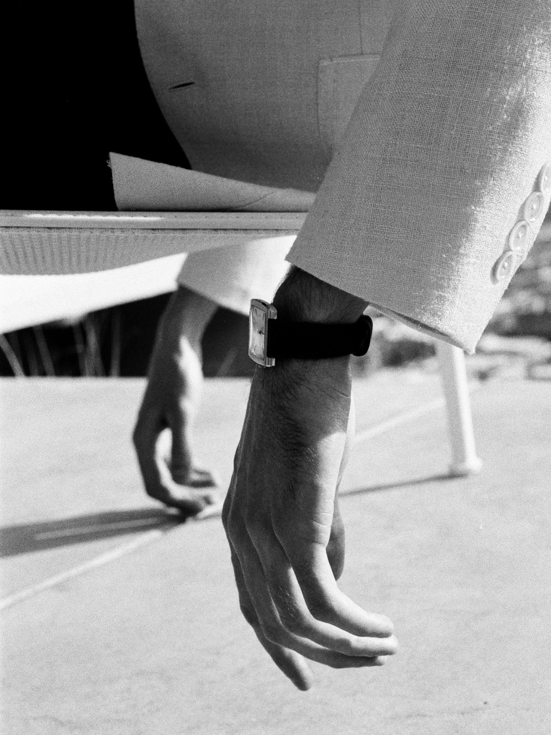 A man's arm with a watch on his wrist hangs down, with his other hand visible in the background.