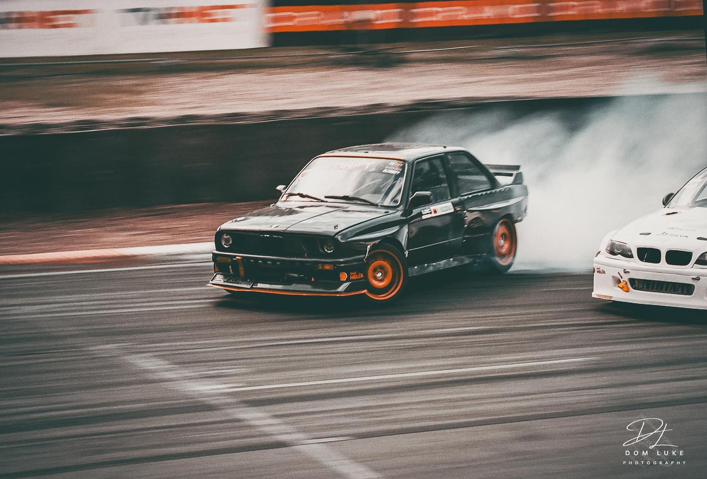 A black car with orange wheels drifts on a race track, creating a cloud of smoke.