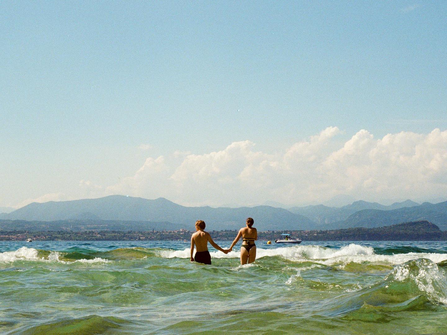 A couple holds hands while standing in the wavy water of a lake with mountains in the background.