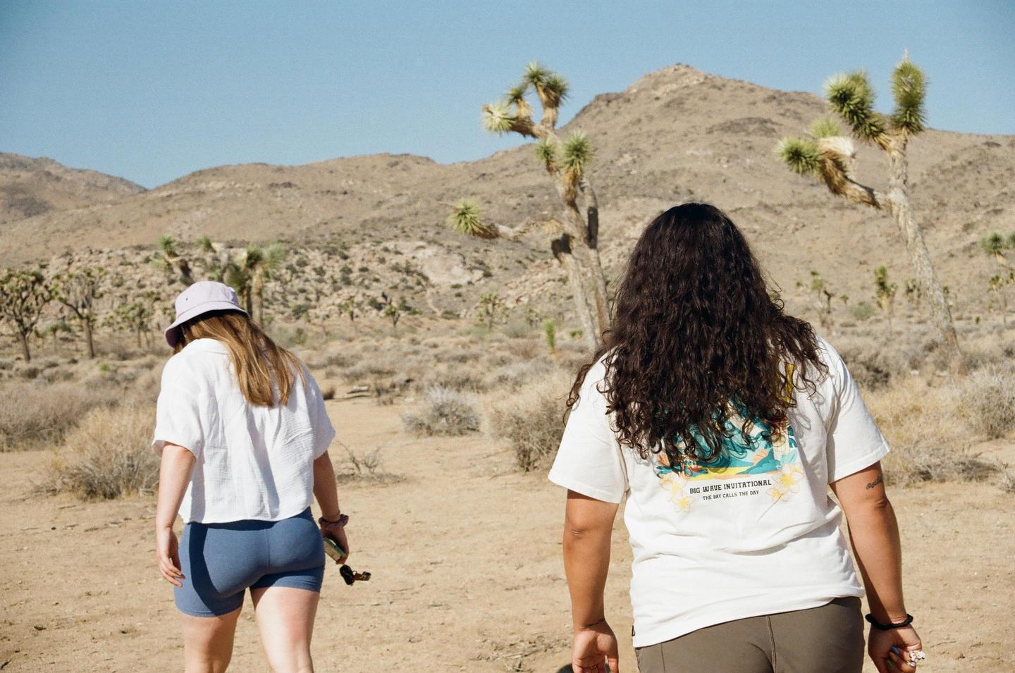 Two women walk away from the camera through a desert landscape with Joshua trees and mountains.