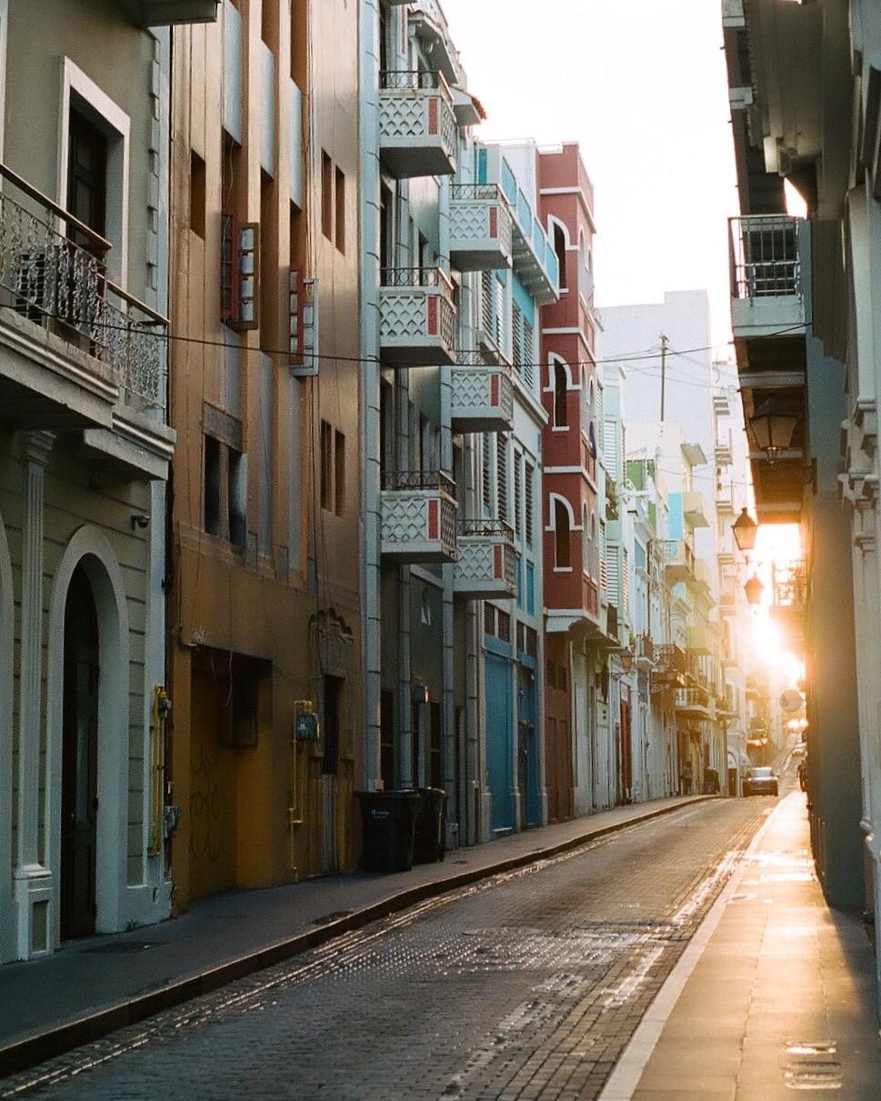 The sun shines brightly down a narrow street lined with colorful buildings and balconies.