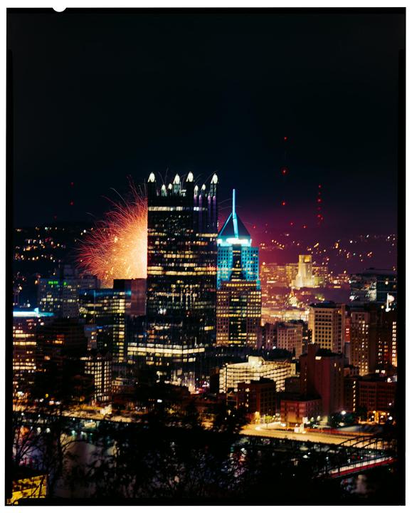 Fireworks explode over a city skyline at night, with illuminated buildings and a bridge visible in the foreground.