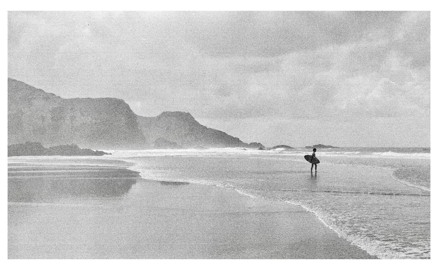 A surfer stands in the shallow water of a beach with mountains in the background.