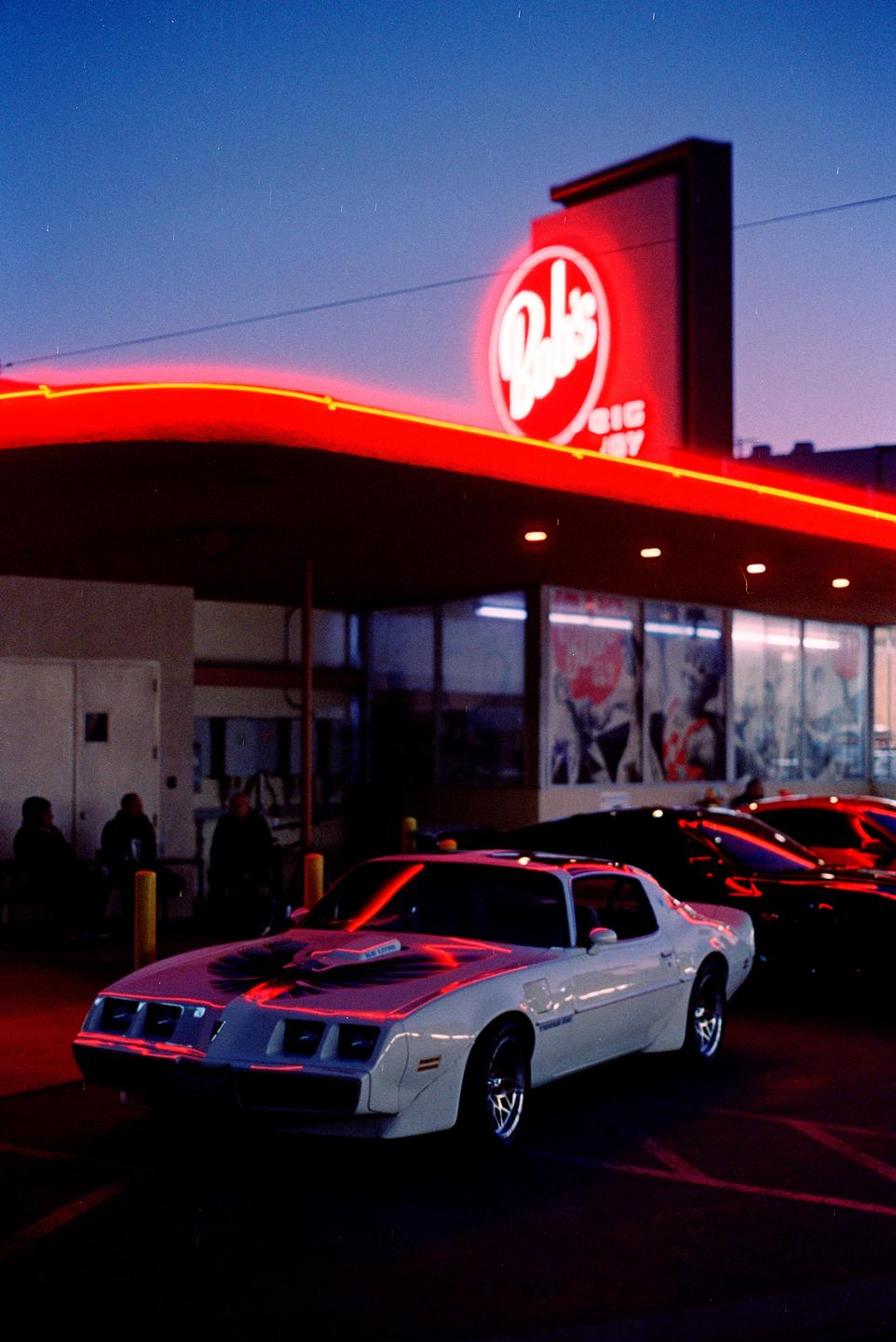 A white car with a bird decal on the hood is parked under a building with a red neon sign at night.