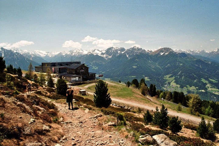 A woman with a walking stick stands on a mountain path overlooking a building and snow-capped mountains.
