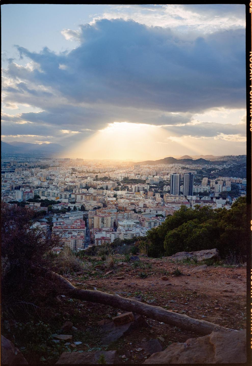 Sunlight streams through clouds over a city with mountains in the background.