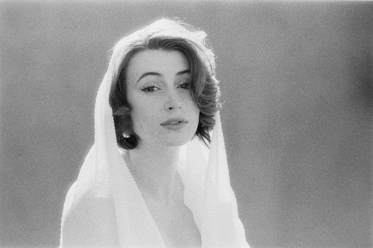 A woman with dark, curly hair and winged eyeliner looks directly at the viewer while wearing a white, textured hood.
