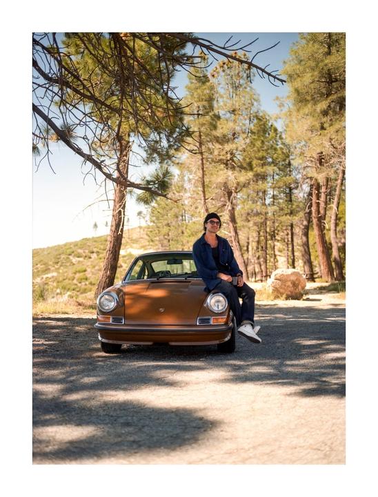 A man sits on the hood of a brown vintage car parked on a dirt road surrounded by pine trees.