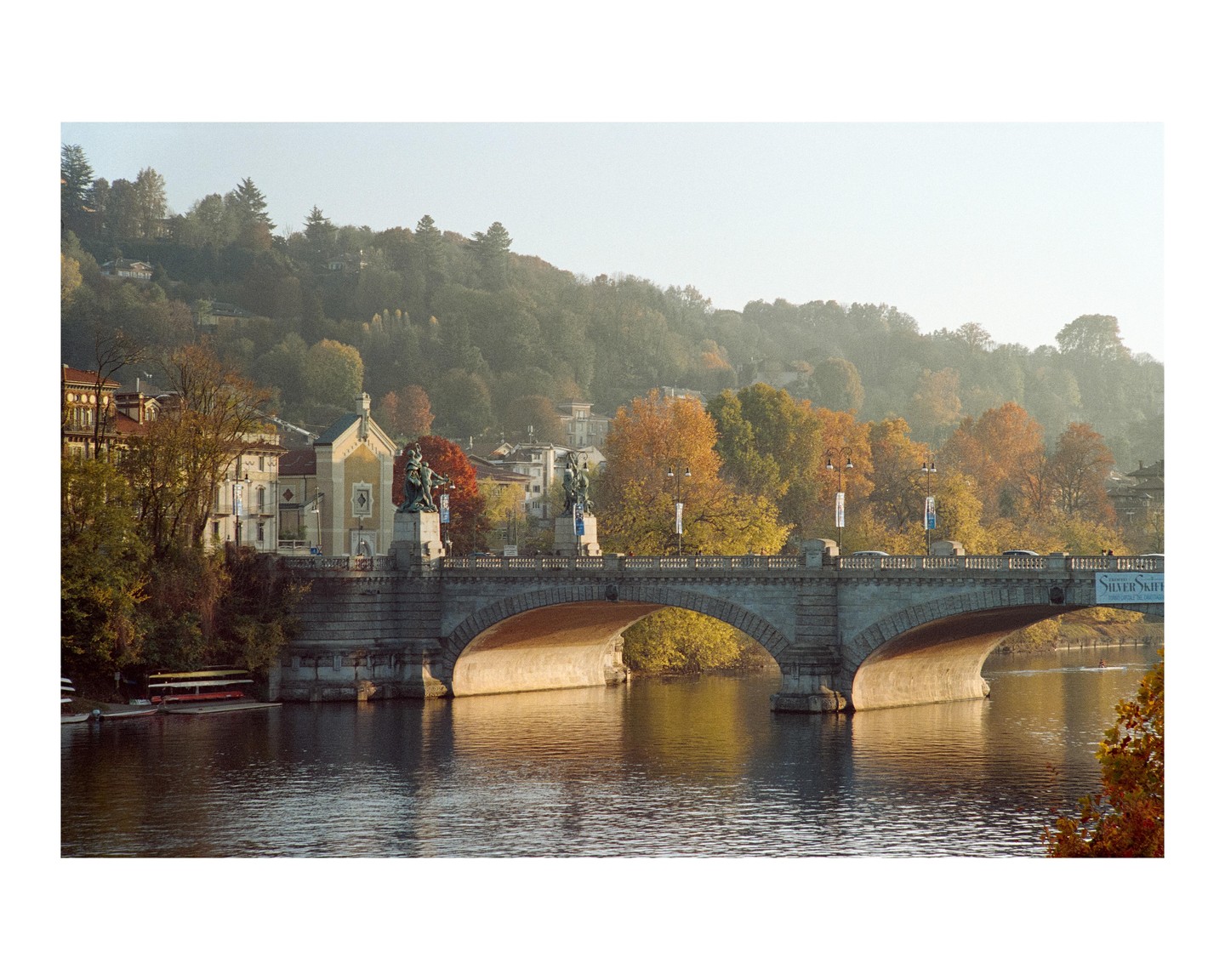 A stone bridge with arches spans a river, with buildings and autumn trees on the far bank and a hazy hillside in the background.