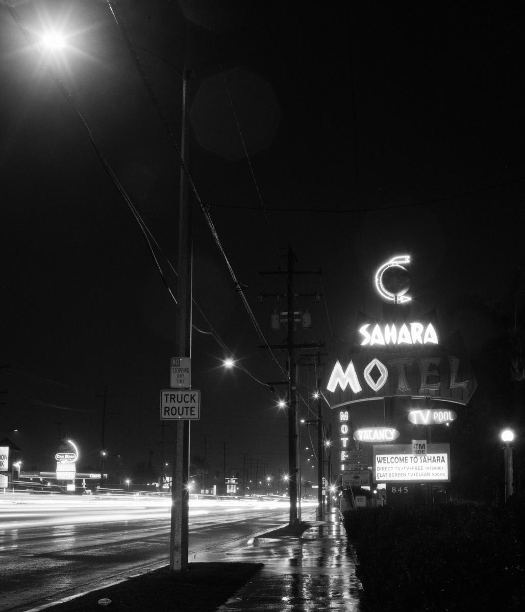 A neon sign for the Sahara Motel stands beside a wet street with light trails from vehicles at night.