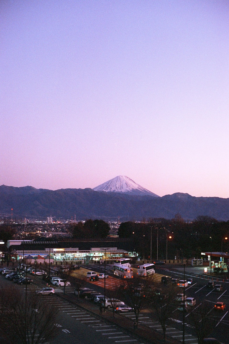 Mount Fuji with a snow-capped peak rises above a town with a parking lot and illuminated buildings at dusk.