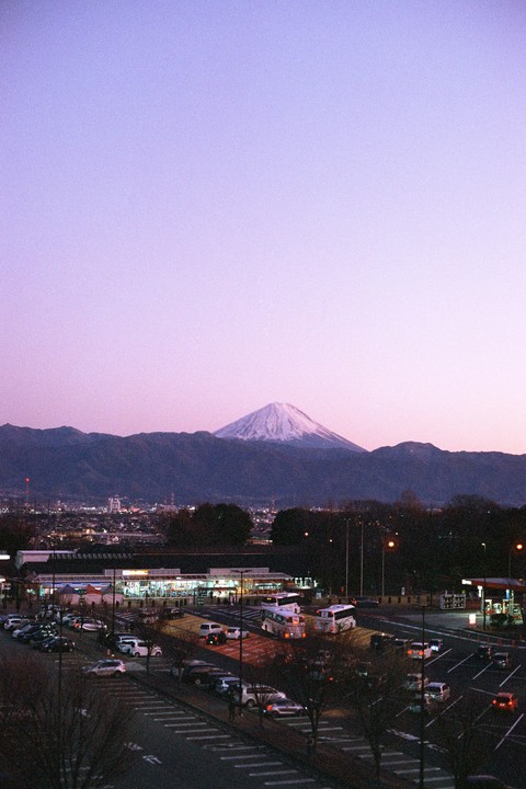 Mount Fuji with a snow-capped peak rises above a town with a parking lot and illuminated buildings at dusk.
