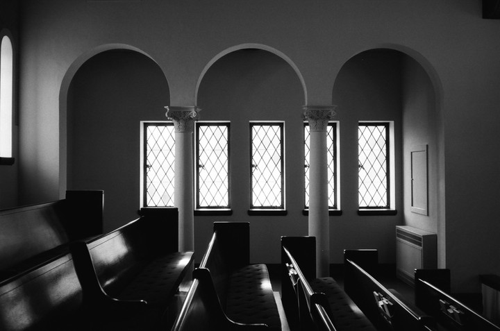 A row of pews faces a wall with arched windows and columns in a church.