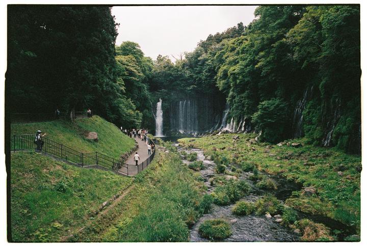 A wide shot shows a waterfall and river surrounded by lush green trees and grass, with a path and people on the left.