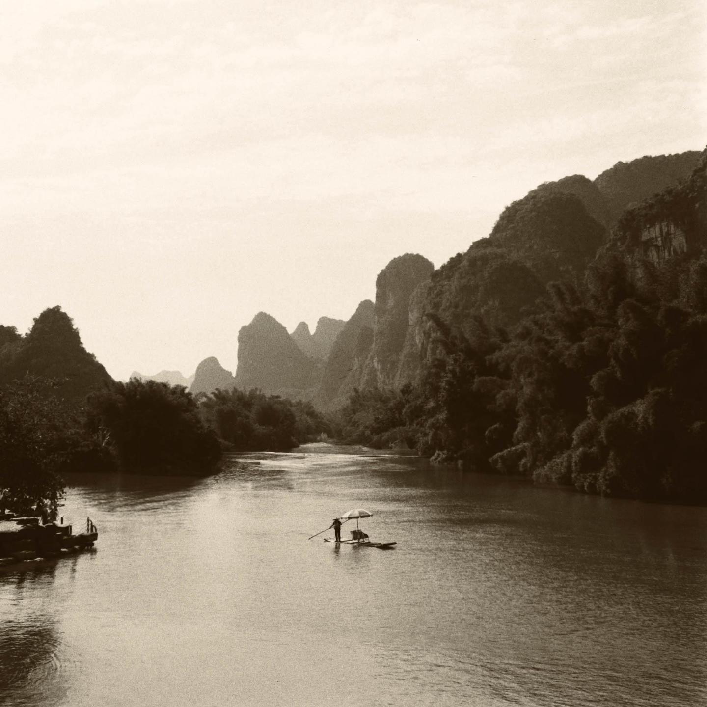 A person punts a small raft with an umbrella on a river surrounded by karstic mountains.