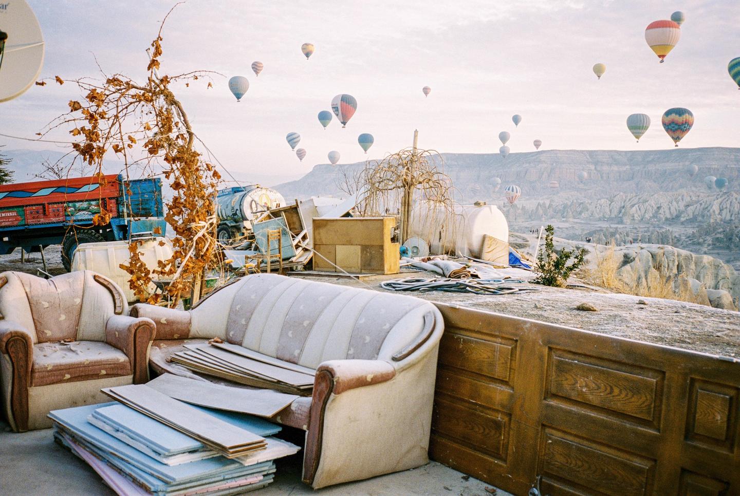 Hot air balloons float over a valley with a truck, furniture, and other items in the foreground.