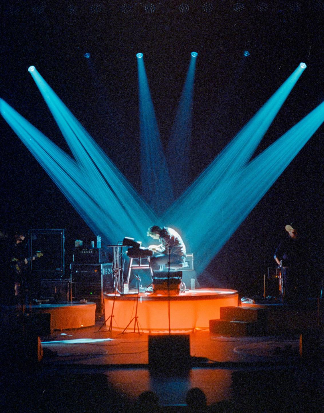 A musician plays a keyboard on a stage illuminated by blue and orange lights.