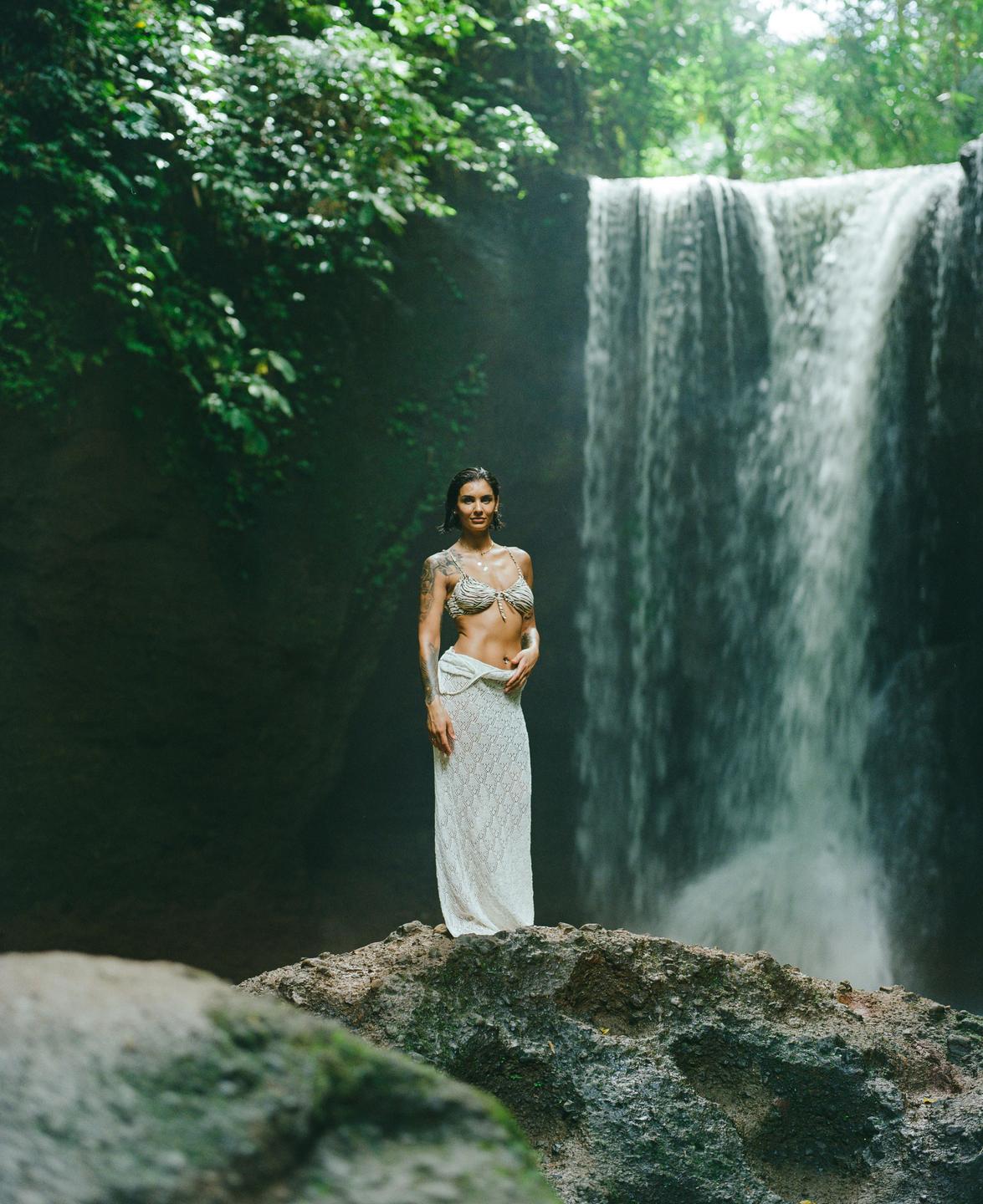 A woman stands on a rock in front of a waterfall in a lush, green environment.