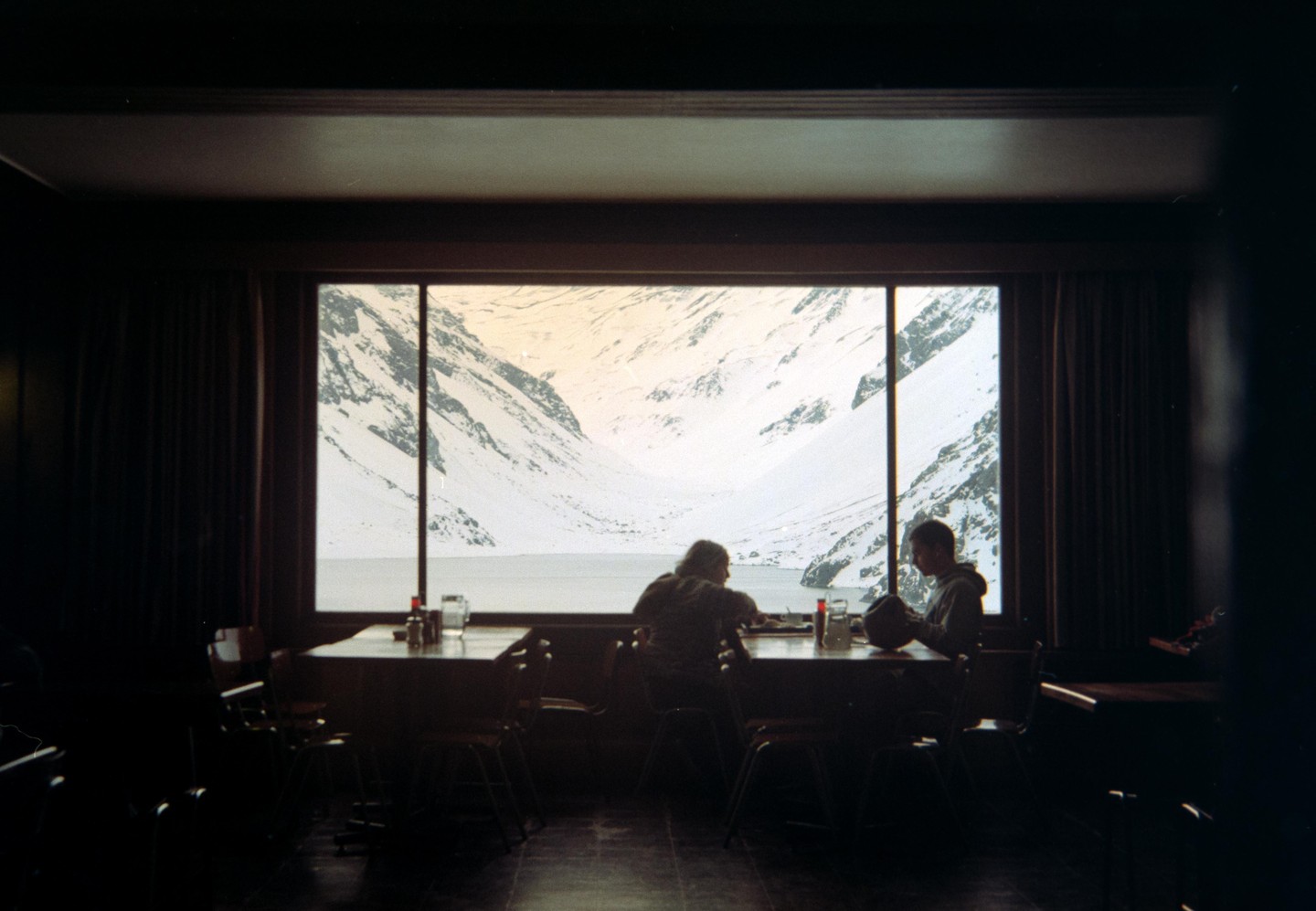 Two people sit at tables in a dark room, looking out a large window at a snow-covered mountain landscape with a body of water.