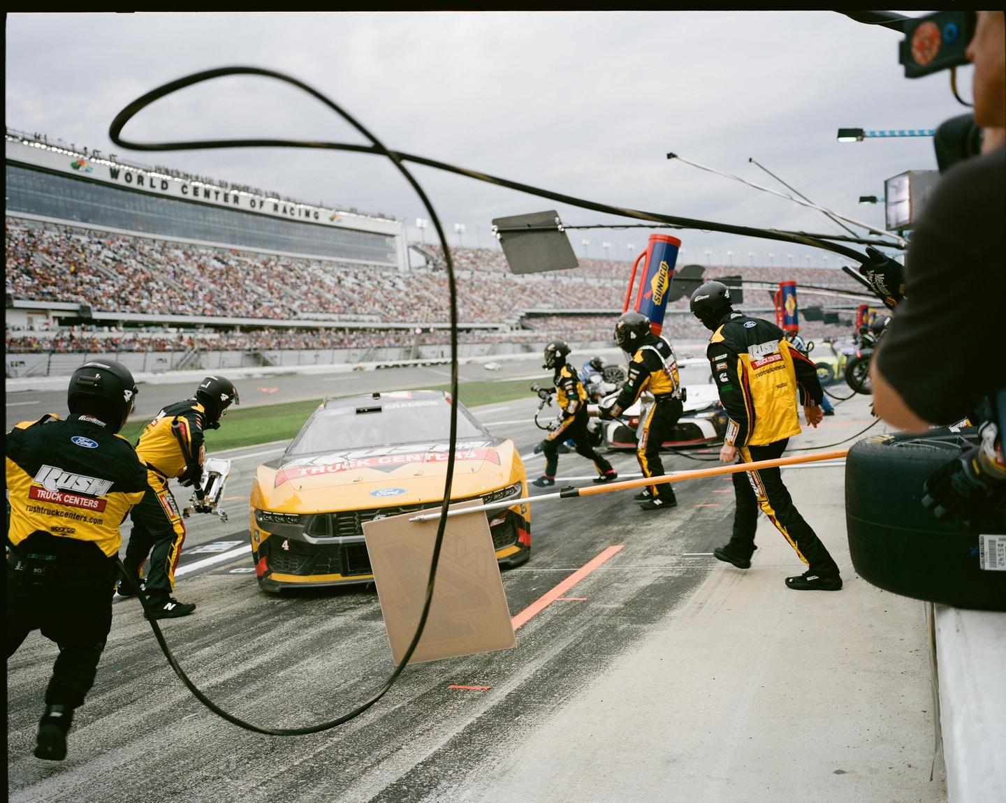 A pit crew services a race car on a track with a grandstand full of spectators in the background.
