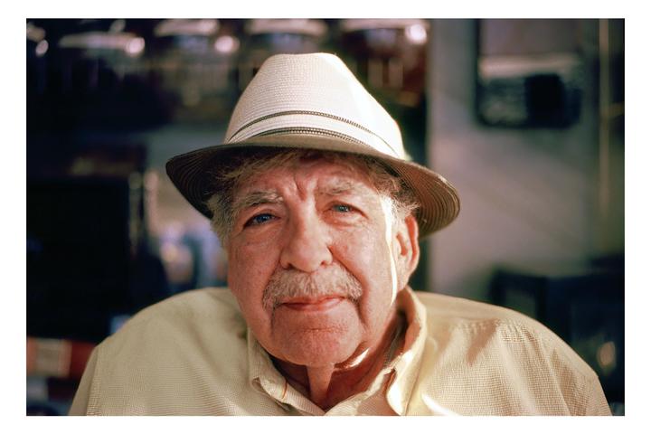 A man wearing a hat and a collared shirt looks directly at the viewer.