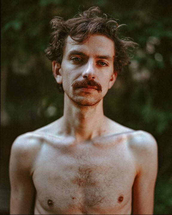 A man with a mustache and curly hair looks directly at the viewer.