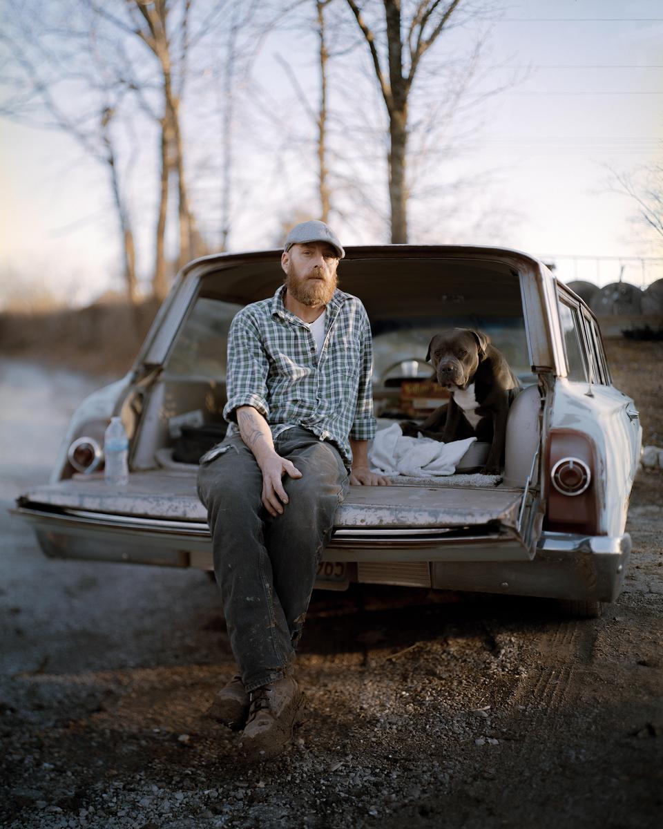 A man with a beard and a dog sit in the open trunk of an old car on a dirt road.