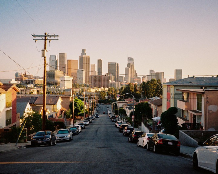 A street with parked cars leads down a hill towards a city skyline with tall buildings.