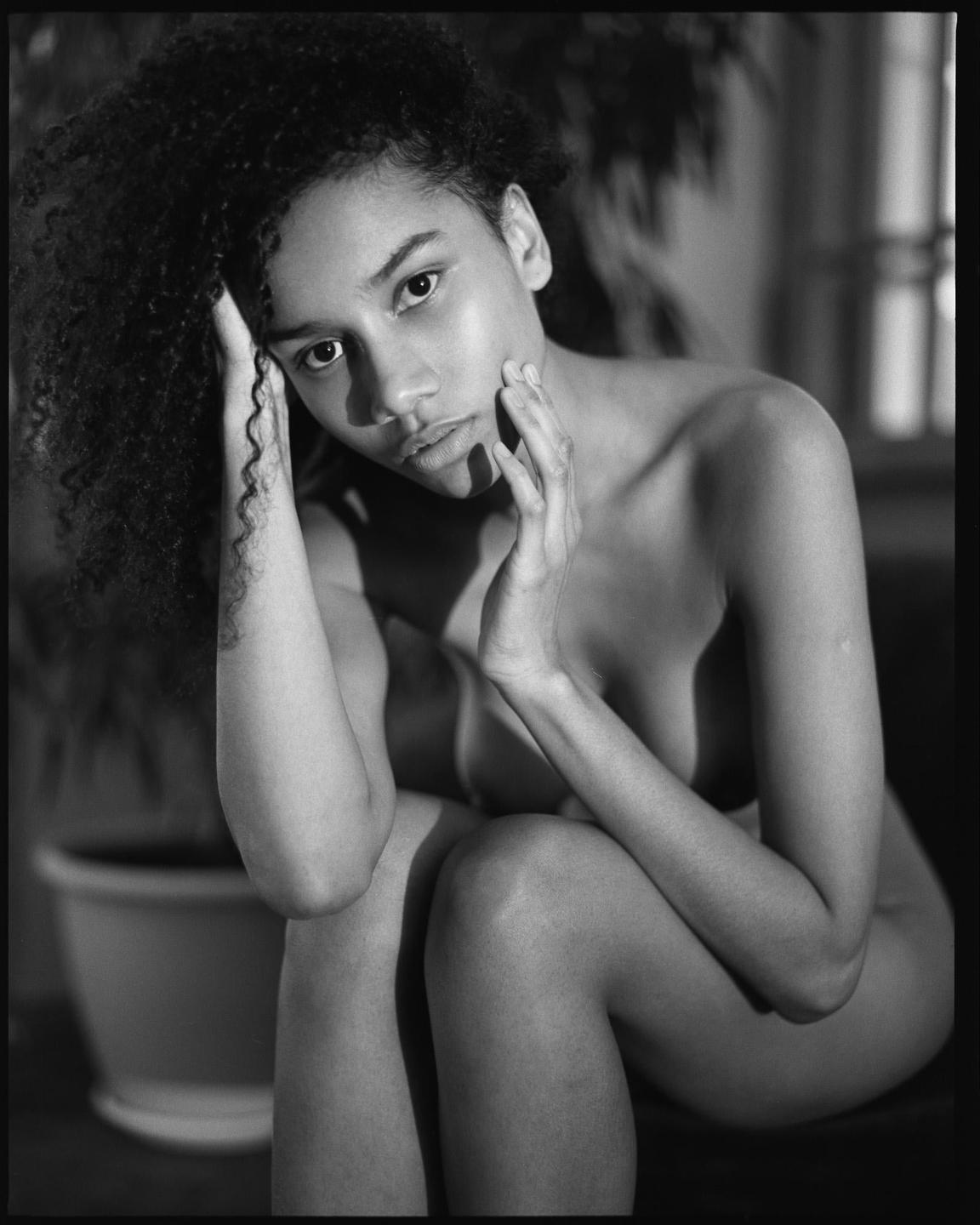 A woman with curly hair sits with her hands near her face, looking directly at the viewer.