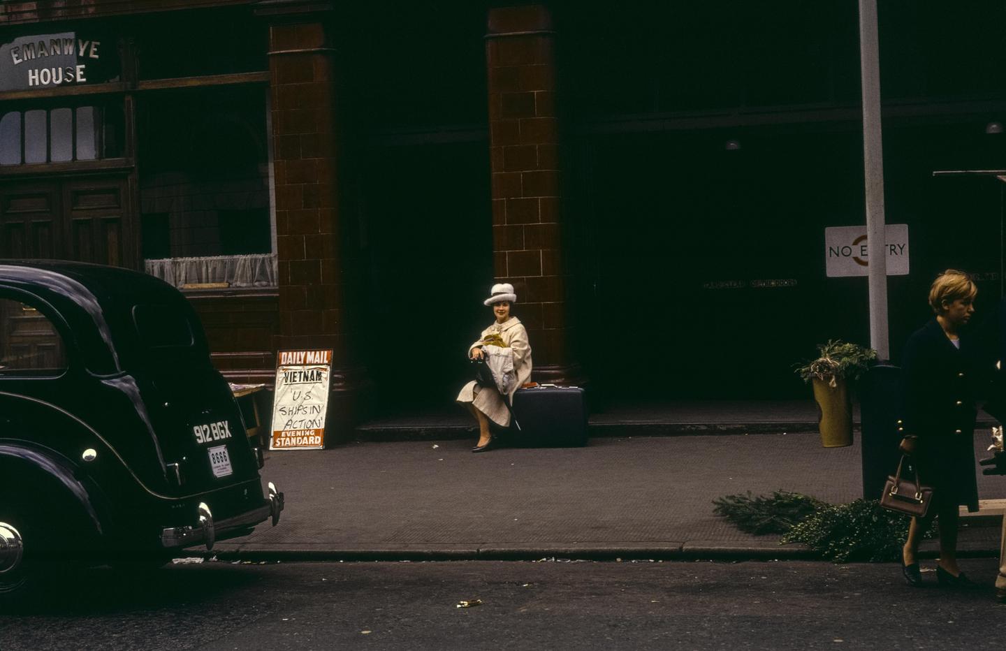 A woman in a hat sits on a suitcase on a city street next to a parked car and a newspaper stand.