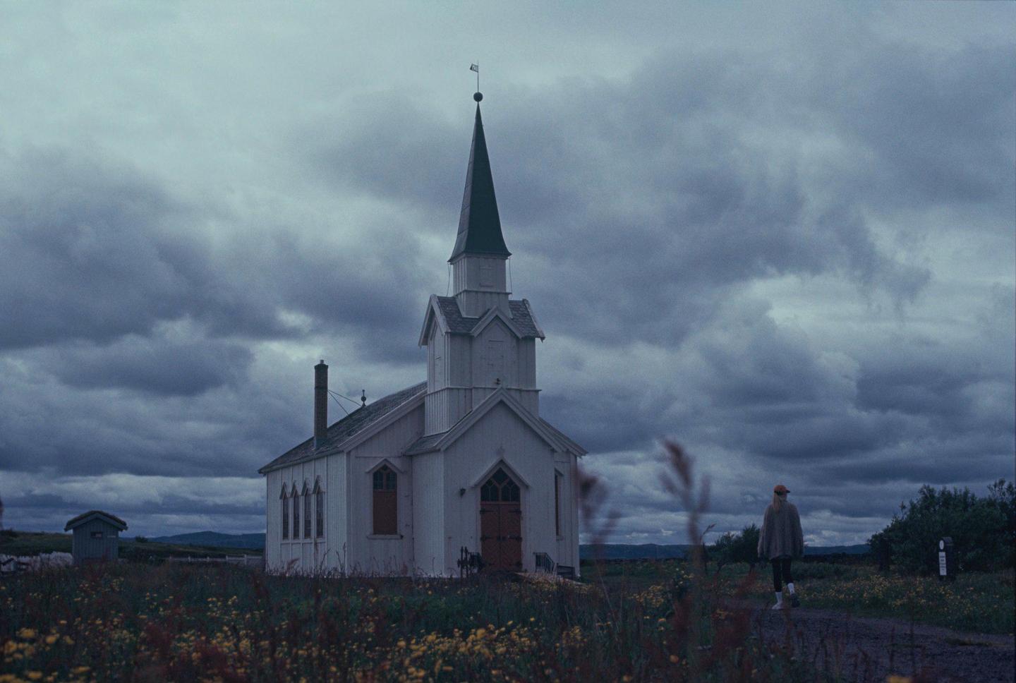 A woman walks on a dirt path next to a white church under a cloudy sky.