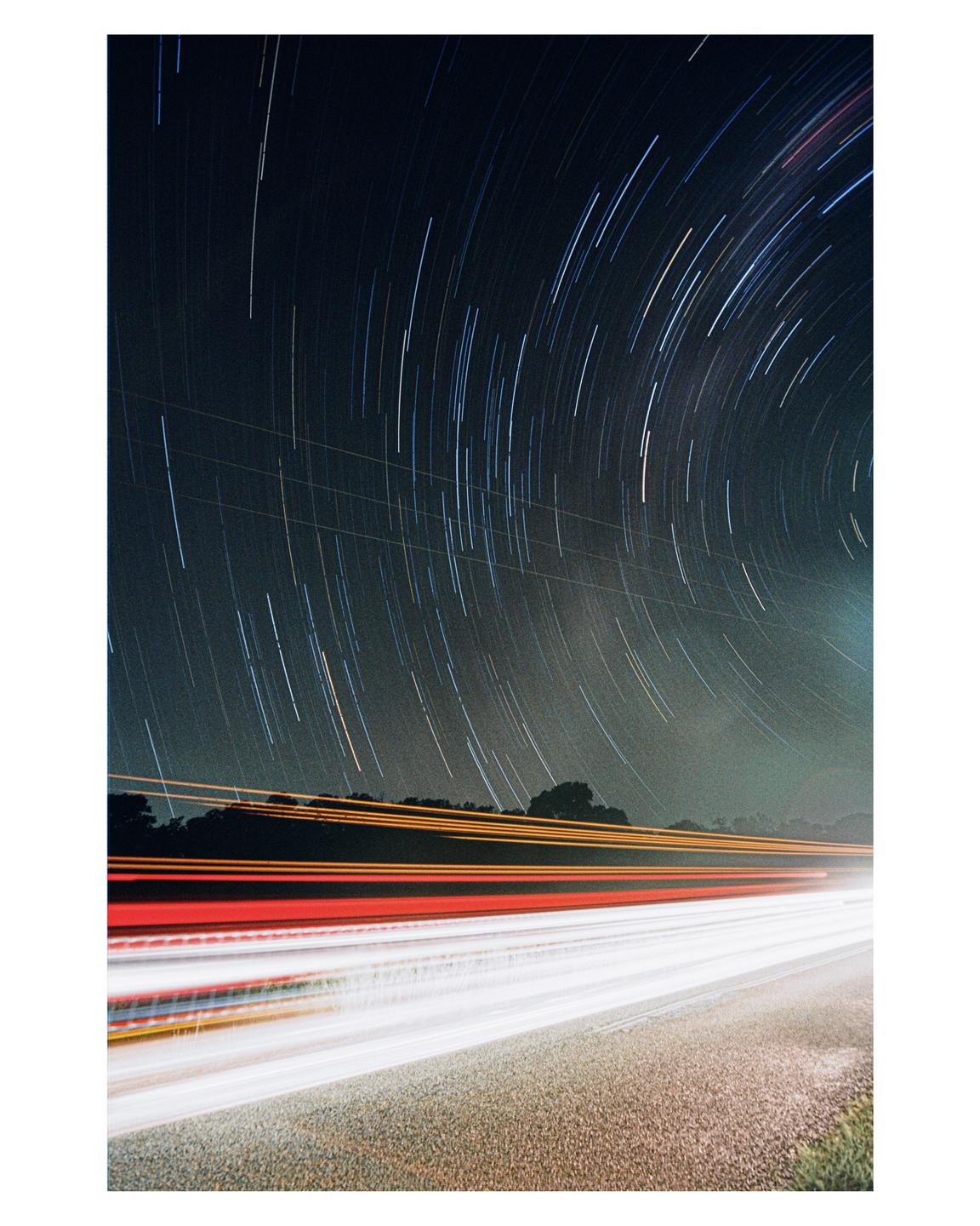 Star trails arc across the night sky above a road with streaks of light from passing vehicles.