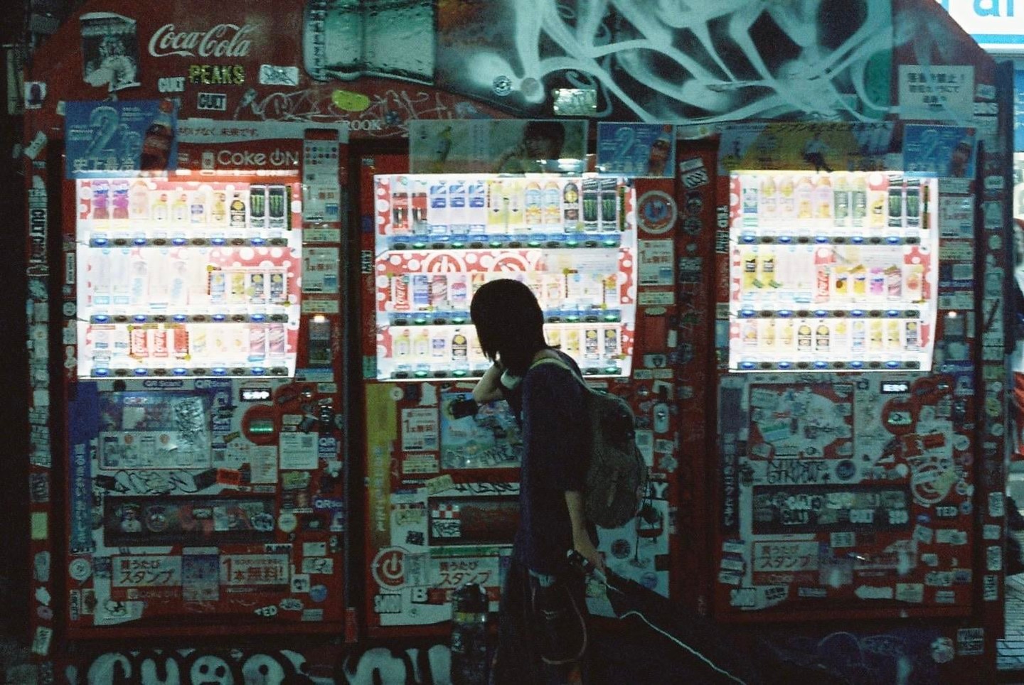 A woman stands in front of three brightly lit vending machines covered in stickers and graffiti at night.