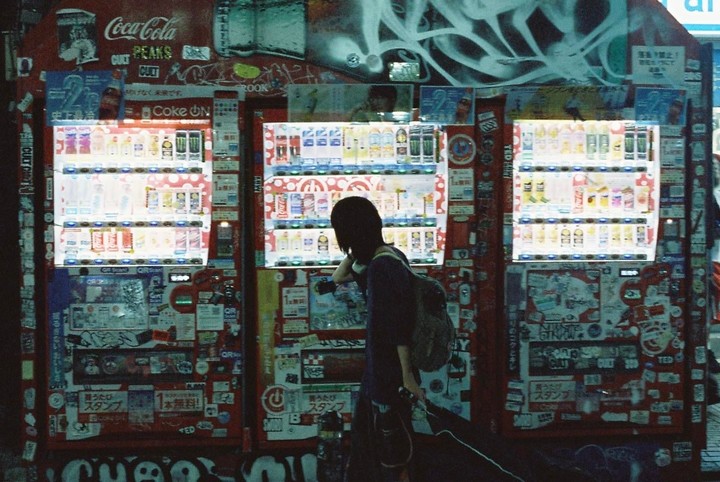 A woman stands in front of three brightly lit vending machines covered in stickers and graffiti at night.