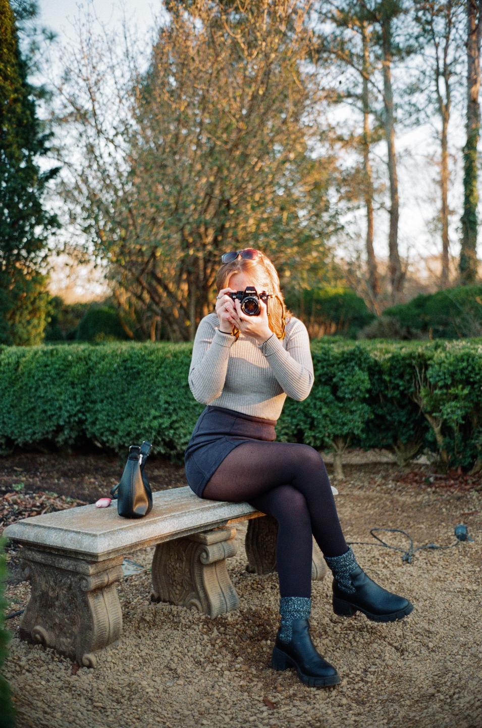 A woman sits on a stone bench and holds a camera to her face, aiming it at the viewer.