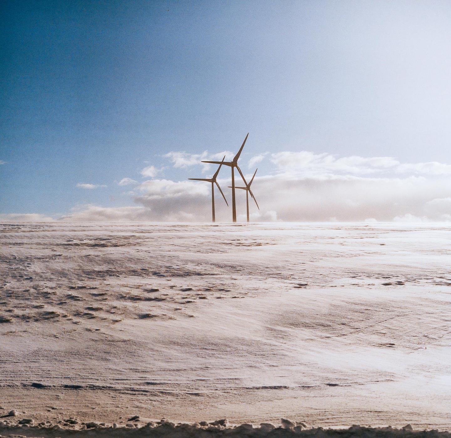 Three wind turbines stand in a snow-covered field under a bright sky with clouds.