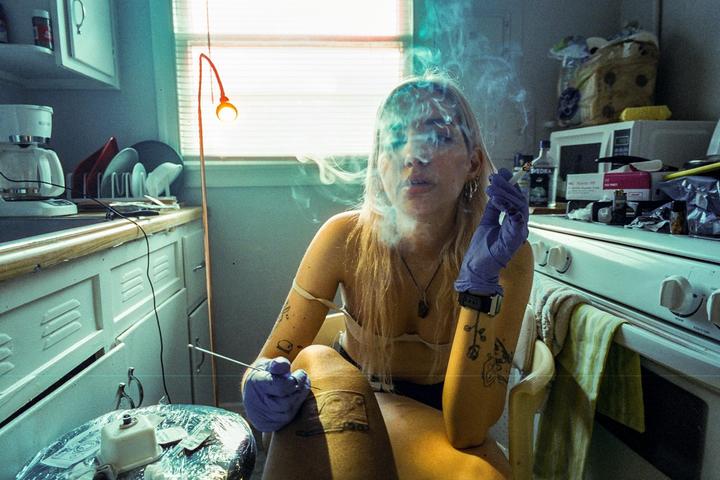 A woman with long hair and tattoos is smoking a cigarette while giving herself a stick and poke tattoo in a kitchen.