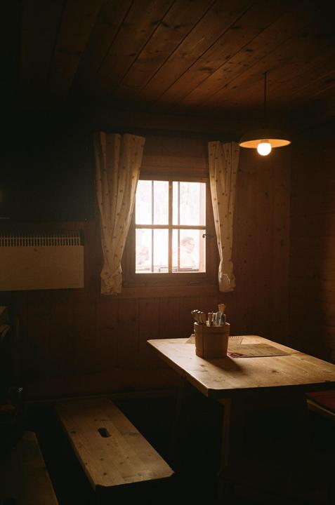 A rustic wooden room features a table with cutlery, a bench, and a window with curtains, through which people are visible.