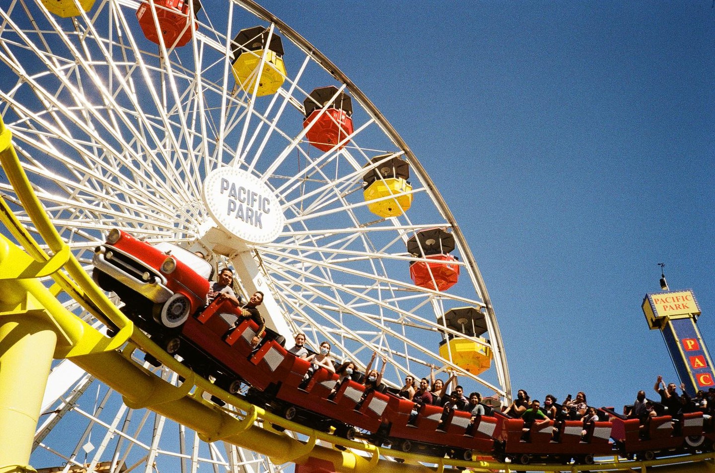 A roller coaster full of people passes in front of a ferris wheel at Pacific Park.