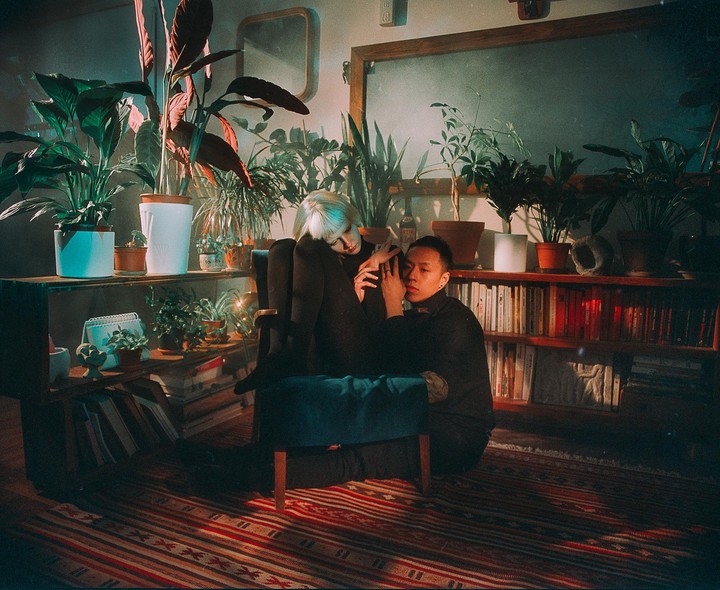 A woman sits in a chair while a man kneels beside her in a room filled with plants and books.