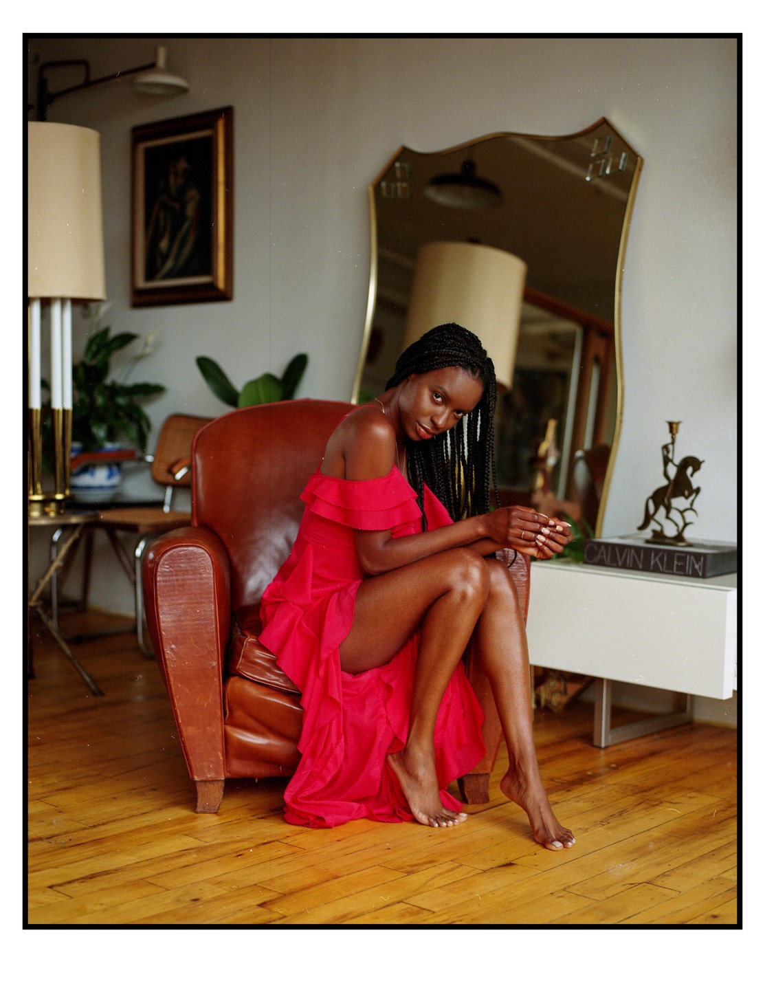 A woman in a red dress sits in a brown leather armchair in a room with a mirror and a lamp.
