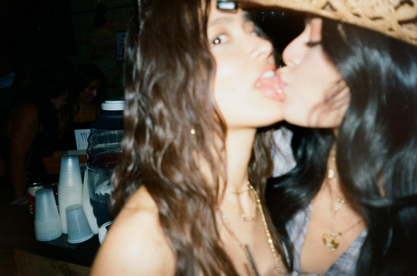 Two women are kissing in a dimly lit room with plastic cups and a large beverage dispenser visible on a counter.