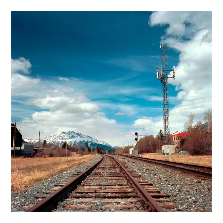 Two parallel railroad tracks curve into the distance toward a snow-capped mountain range under a blue sky with white clouds.